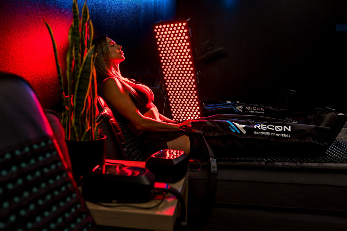 Woman relaxing in a recliner wearing RECON compression boots beside a glowing Restore red light panel