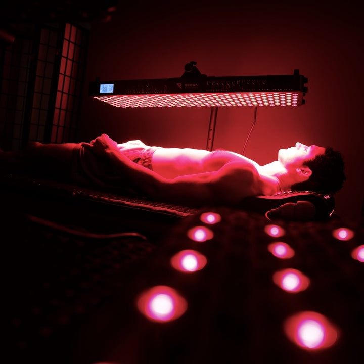 Person lying on a mat receiving red light therapy in a dimly lit room.