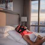 Woman lying on a bed wearing a red light therapy mask with a cityscape .