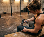 Athlete seated on a box using the RECON Pulse Massage Gun on her calf in a gym